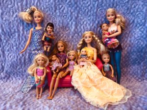 A picture of 11 Barbie Dolls against a purple background for a Barbie Doll Story.  It's set up like a family portrait.  3 Actual Barbie Dolls, 1 Stacie, 3 Chelsea Dolls, 2 toddler dolls and 2 baby dolls. 