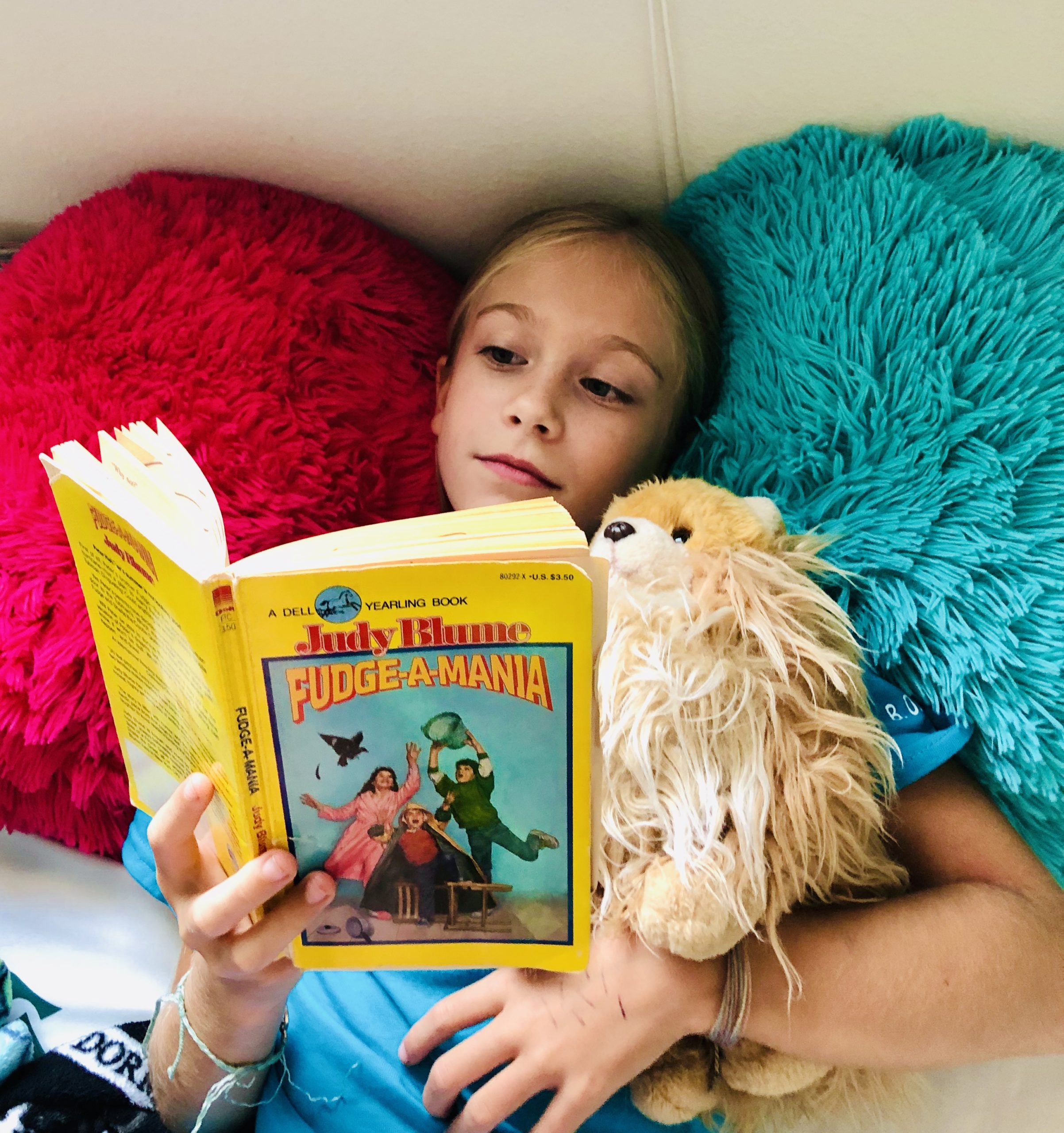 For the book review, a blond haired girl, Brecken, reading Fudge-A-Mania by Judy Blume. She's laying on 2 fuzzy pillows, hot pink and blue. 