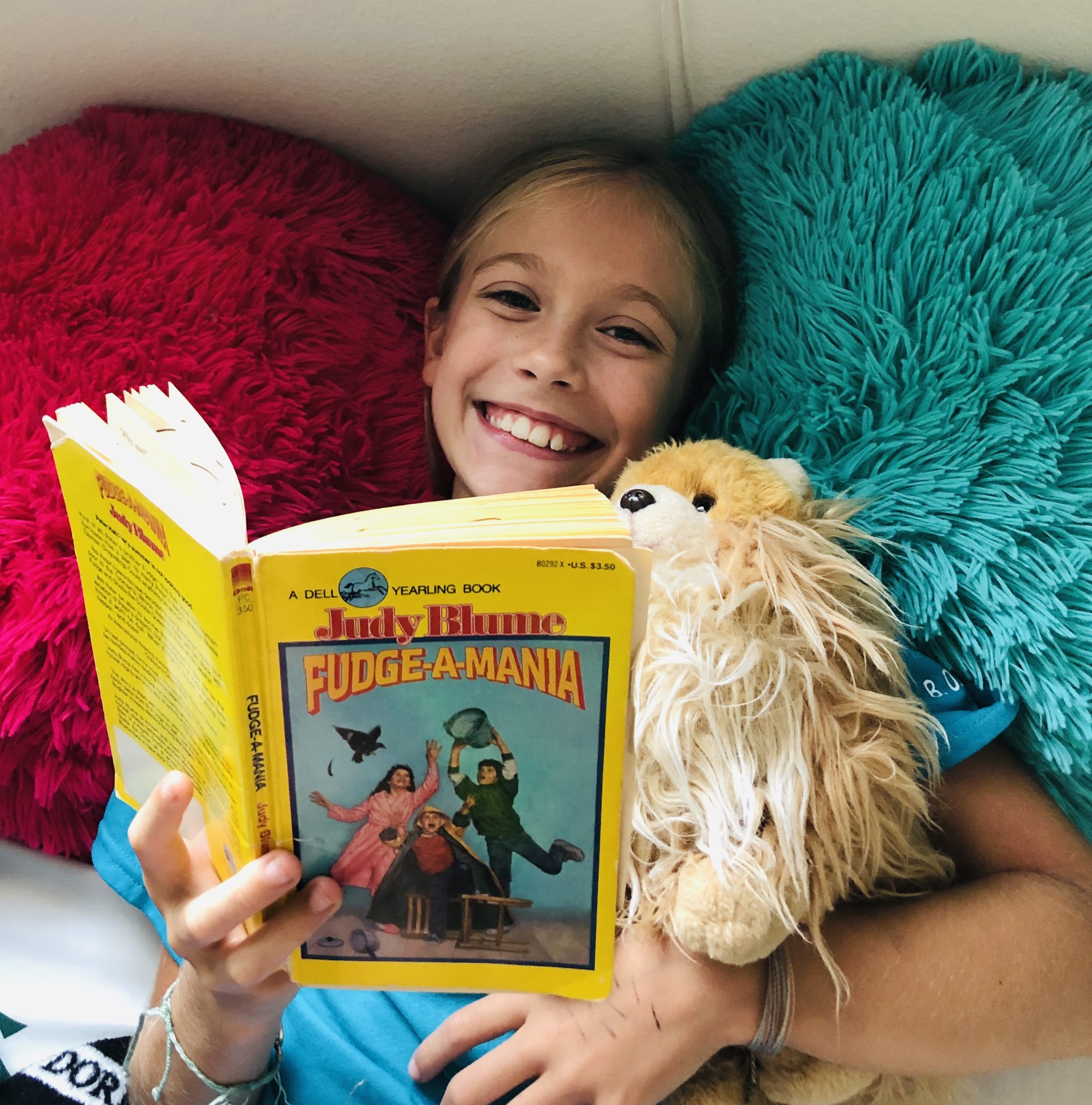 For the book review, a blond haired girl, Brecken, reading Fudge-A-Mania by Judy Blume. She's laying on 2 fuzzy pillows, hot pink and blue. She's smiling.