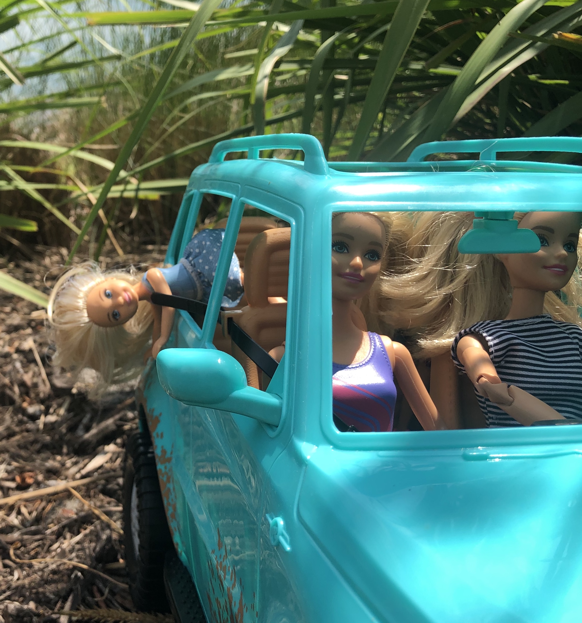 A Chelsea doll falling out the backseat of a teal Jeep. Her head and upper body is hanging down.