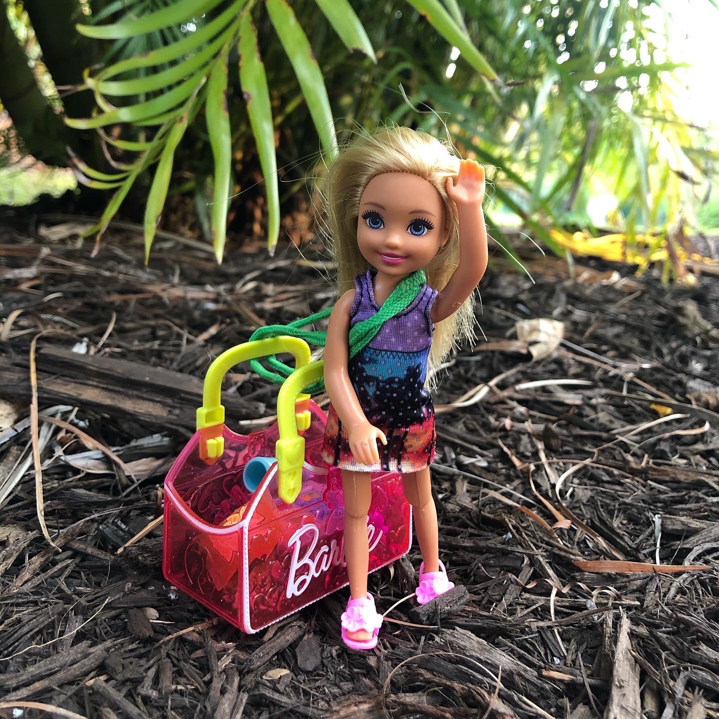 A Chelsea doll waving at the camera with a pink Barbie bag next to her for the Barbie story for kids, Hiking in Costa Rica.