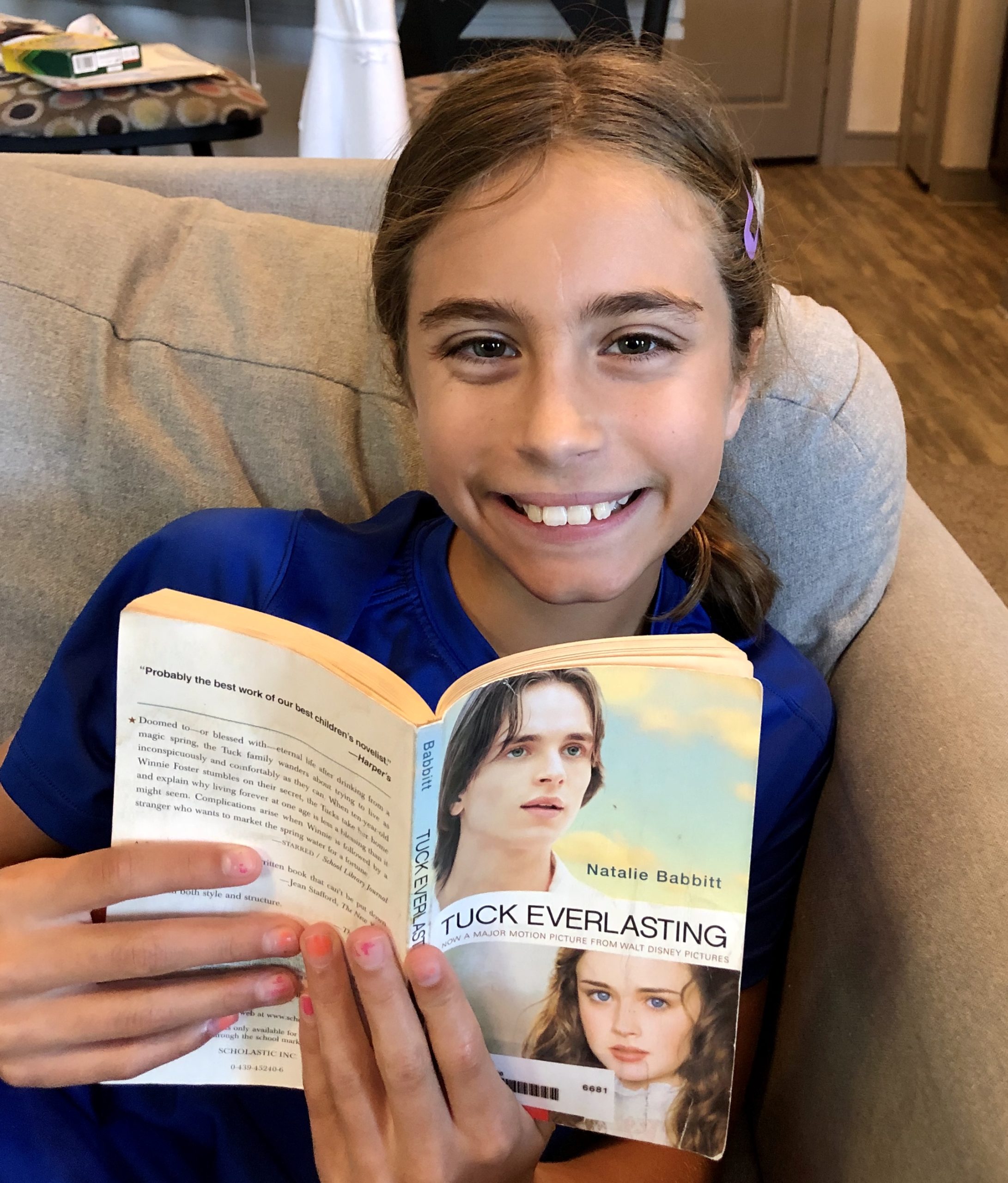 A young girl smiling at the camera, while holding the book Tuck Everlasting by Natalie Babbitt for a children's Book Review by Kandblife.com