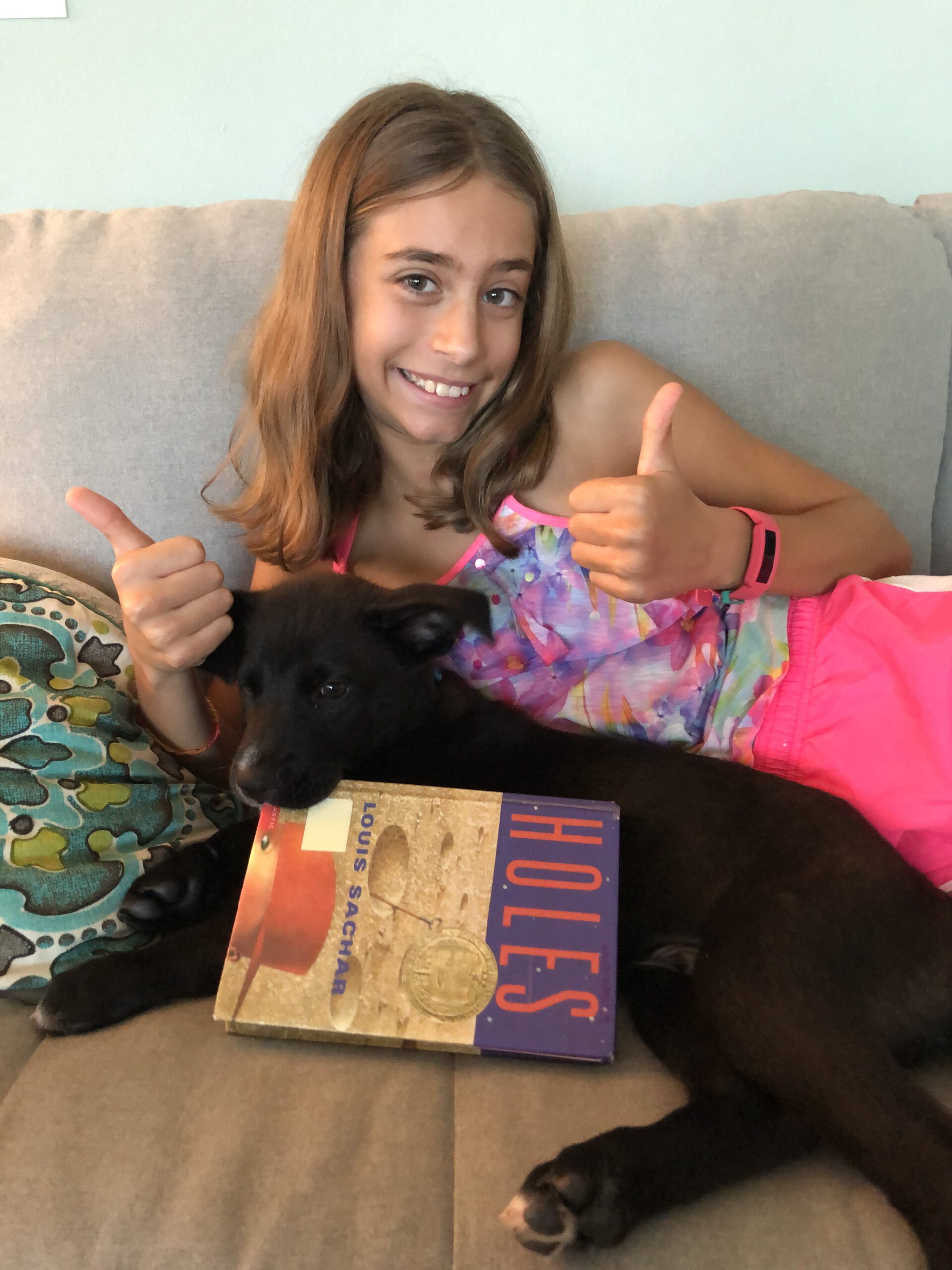 Girl, laying with a black dog, holding her thumbs up for the book review on Holes.